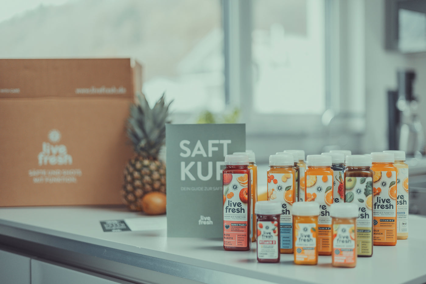 Verschiedene bunte Saftflaschen und Shots der Marke LiveFresh stehen auf einer hellen Küchenarbeitsfläche neben einer grünen Broschüre mit der Aufschrift Saftkur, einer Ananas und einem Versandkarton.