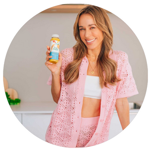 Eine lächelnde Frau mit langen, welligen Haaren trägt ein rosa Strickset und hält eine Flasche „Live Fresh“-Saft in der Hand. Sie steht in einer modernen Küche mit einer Reihe bunter Säfte vor sich. Im Vordergrund ist ein grünes Schild mit der Aufschrift „SAFT KUR“ zu sehen.