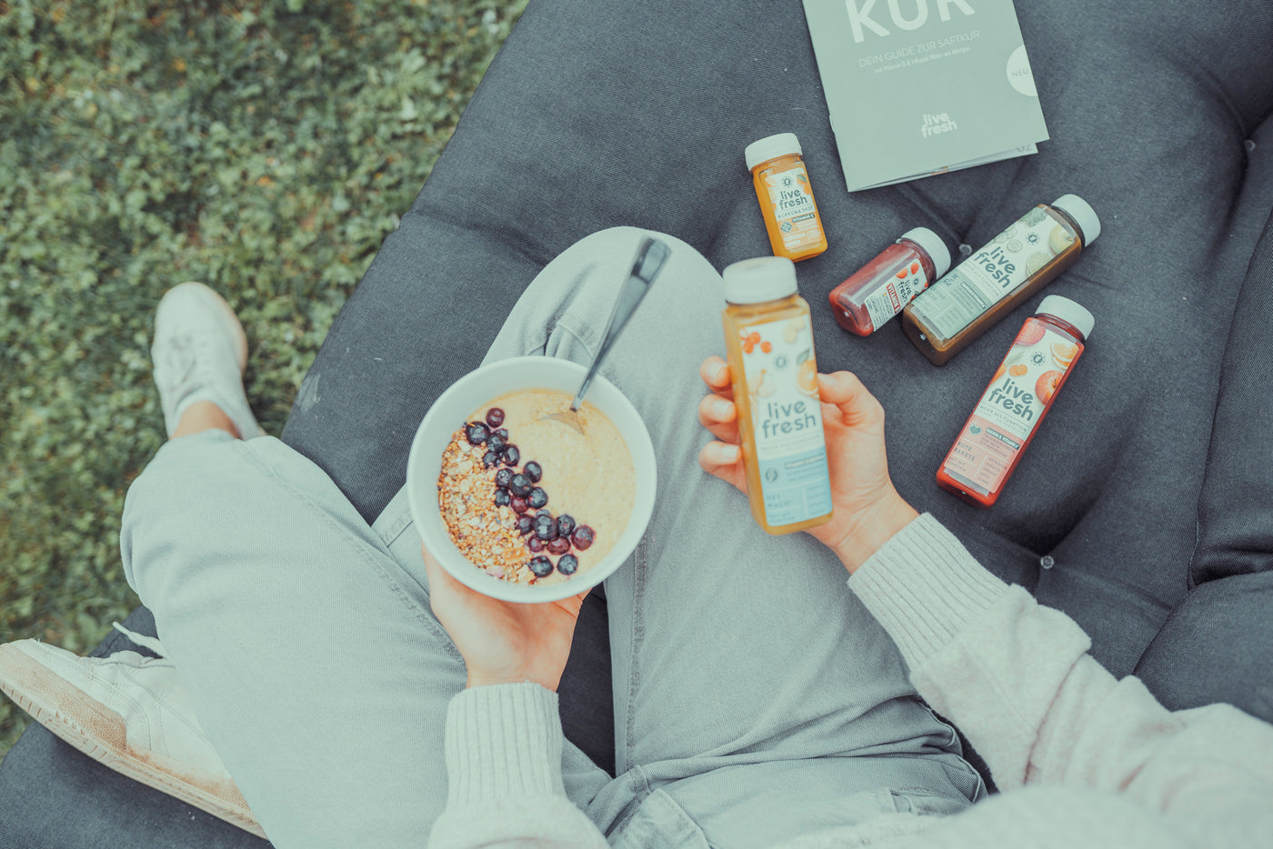 Eine Person sitzt entspannt auf einer dunklen Outdoor-Couch mit gekreuzten Beinen, trägt helle Kleidung und weiße Sneaker. In einer Hand hält sie eine Schüssel mit Porridge, Granola und Heidelbeeren, in der anderen eine Flasche LiveFresh Saft „Hey Wach+“. Mehrere weitere LiveFresh Säfte und ein Saftkur-Guide liegen neben ihr auf der Couch. Im Hintergrund ist eine grüne Wiese zu sehen.