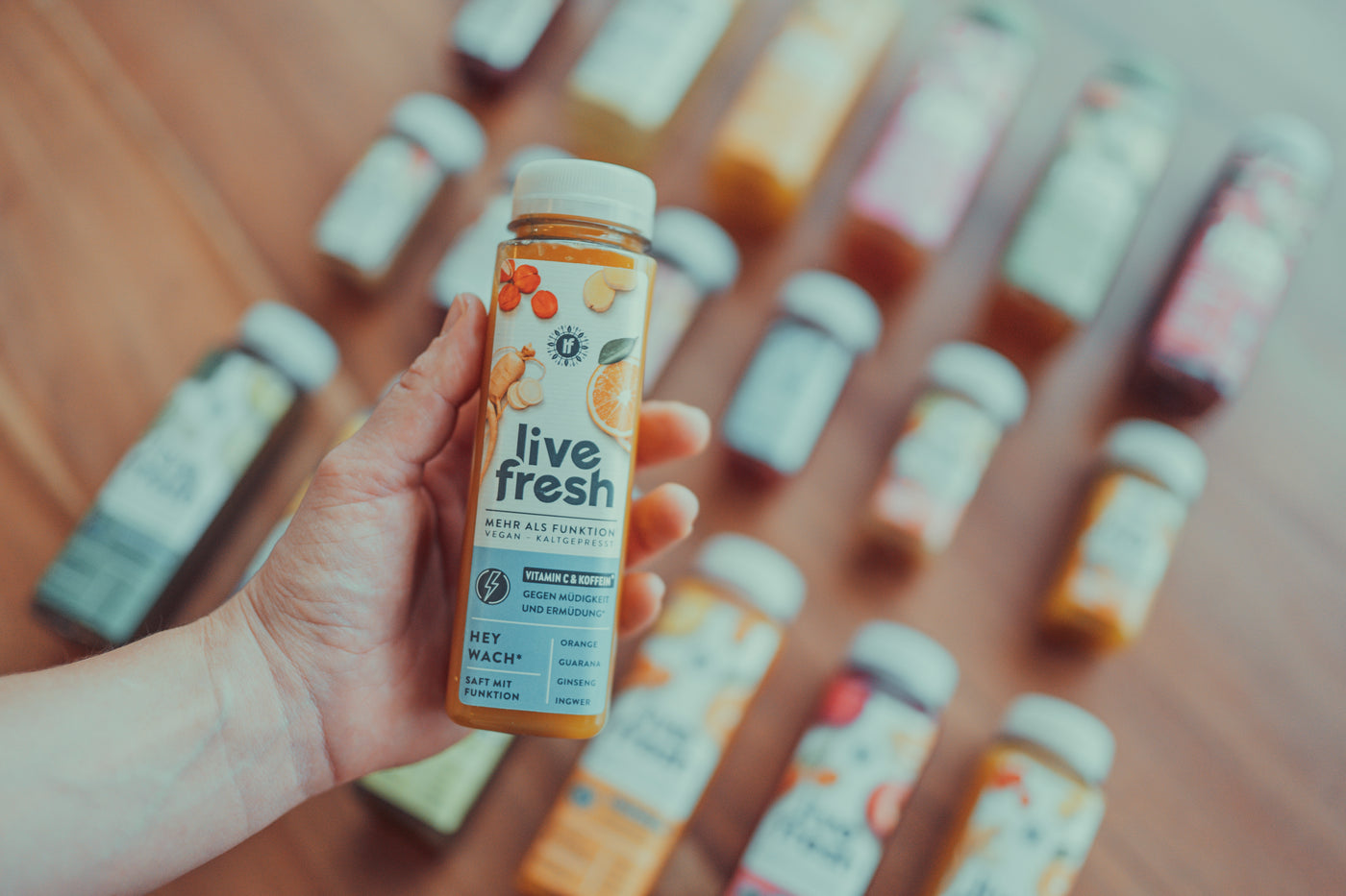 Eine Hand hält eine Flasche LiveFresh Saft „Hey Wach+“ mit Vitamin C und Koffein, hergestellt aus Orange, Guarana, Ginseng und Ingwer. Im Hintergrund liegen weitere LiveFresh Säfte in verschiedenen Sorten, leicht unscharf auf einer Holzoberfläche.