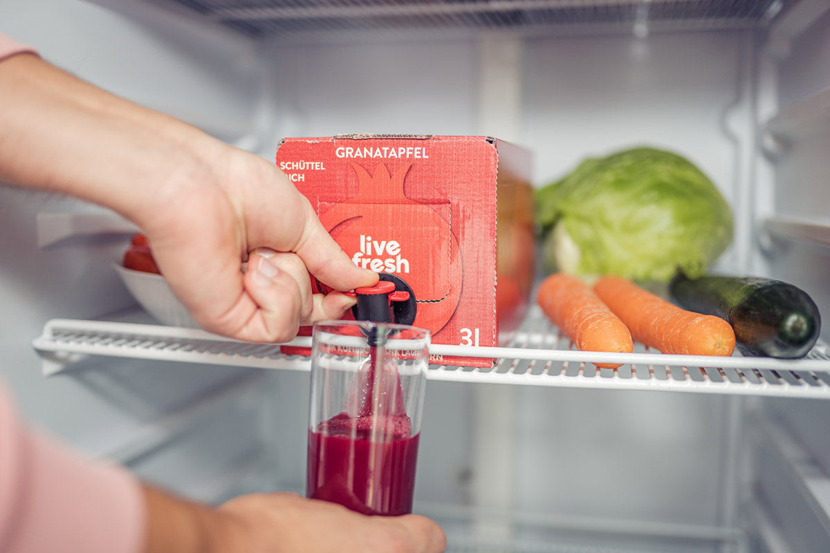 Eine Hand, die in einem Kühlschrank ein Glas mit Granatapfelsaft aus einer lachsfarbenen„LiveFresh“ 3-Liter-Box mit kaltgepresstem Saft zapft. Auf der Saftbox steht „Granatapfel – Schüttel mich“. Neben der Box liegen frische Karotten, Salat und eine Gurke. Der Zapfhahn der Box wird betätigt, um den frischen Granatapfelsaft ins Glas zu füllen.