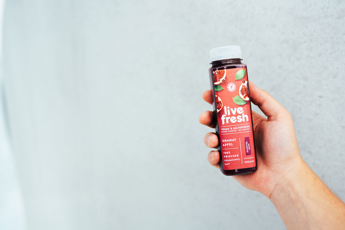 Eine Hand, die eine Flasche „LiveFresh“ Granatapfelsaft hält. Auf dem roten Etikett steht „Vegan & Kaltgepresst“, „100% Frischer Granatapfelsaft“, sowie „Keine Zusätze – Nie Erhitzt“. Die Flasche ist vor einem grauen, hellen Hintergrund abgebildet.