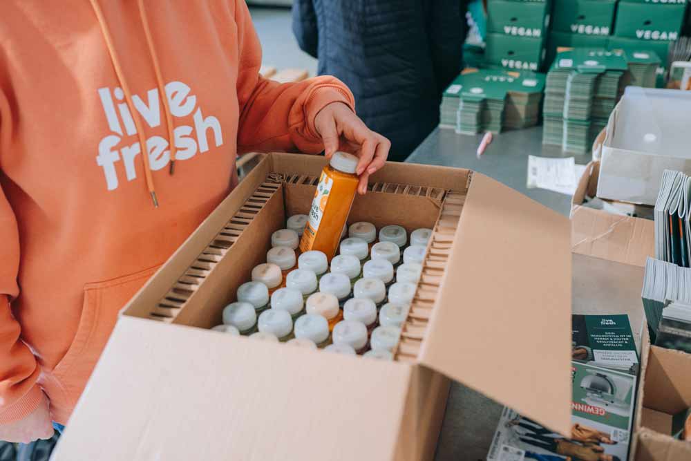 Eine Person in einem orangefarbenen „LiveFresh“-Hoodie verpackt kleine Saftflaschen in einen Karton. Die Flaschen sind ordentlich angeordnet, eine orangene wird gerade in die Hand genommen. Im Hintergrund liegen grüne Verpackungen mit der Aufschrift „VEGAN“ sowie Werbematerialien auf dem Tisch.