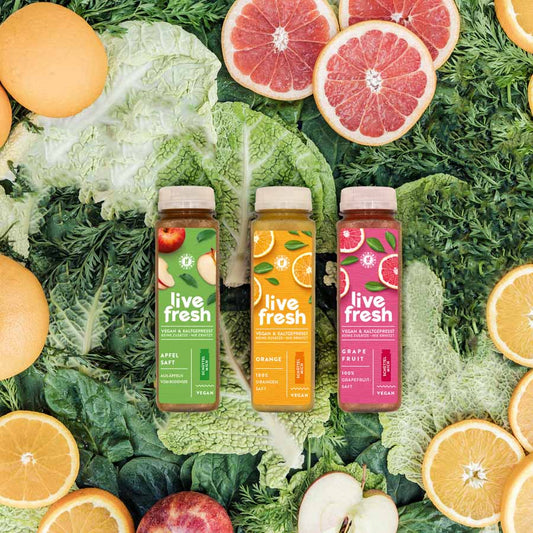Drei Flaschen LiveFresh Saft stehen nebeneinander auf einem Hintergrund aus grünem Gemüse und aufgeschnittenen Orangen-, Grapefruit- und Apfelscheiben. Die Sorten umfassen Apfelsaft (grünes Etikett), Orangensaft (gelbes Etikett) und Grapefruitsaft (pinkes Etikett), alle kaltgepresst und vegan.