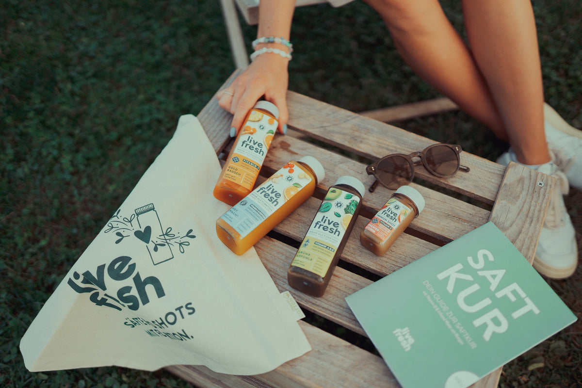 Vier “Live Fresh”-Saftflaschen in verschiedenen Sorten auf einer Holzbank, daneben eine Stofftasche mit Logo, eine Sonnenbrille, ein hellgrünes Booklet mit dem Titel “SAFT KUR” und ein Teil einer Person mit weißen Sneakers im Hintergrund.