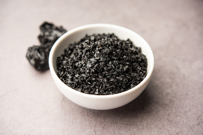 Schale voll mit dunklen Shilajit-Kristallen, auch bekannt als Mumijo, ein natürliches Harz, das in der ayurvedischen Medizin verwendet wird, auf einem grauen Hintergrund.