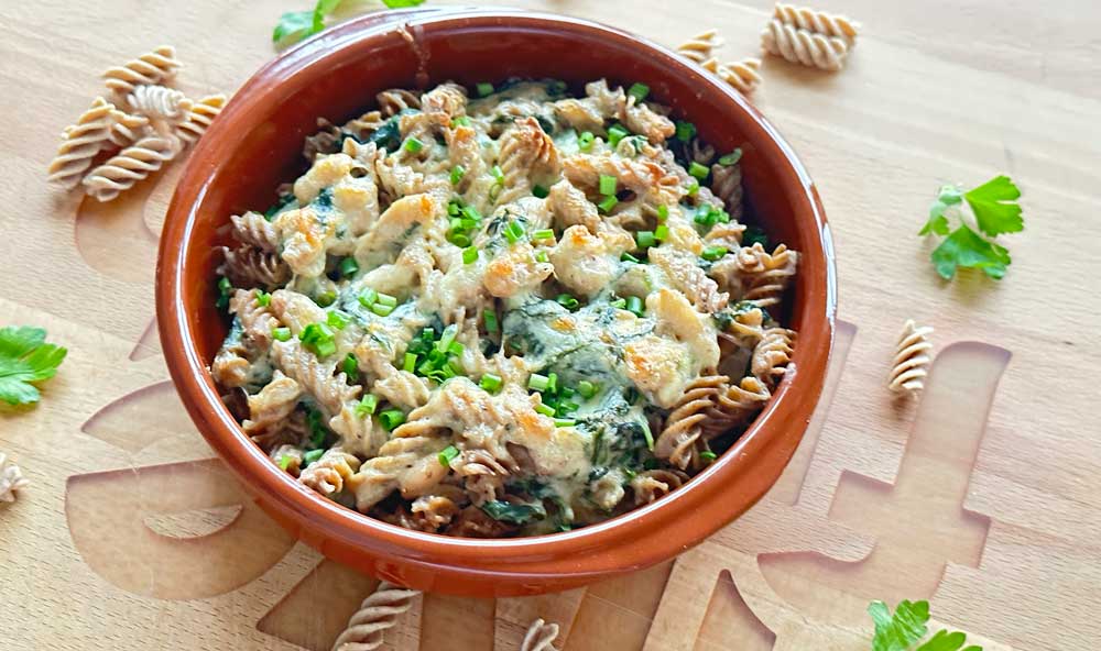 Überbackener Low Carb High Protein Nudelauflauf mit Vollkornfusilli, Spinat und cremiger Käsesauce, garniert mit frisch gehackten Kräutern, serviert in einer terrakottafarbenen Schale auf einem Holzbrett.