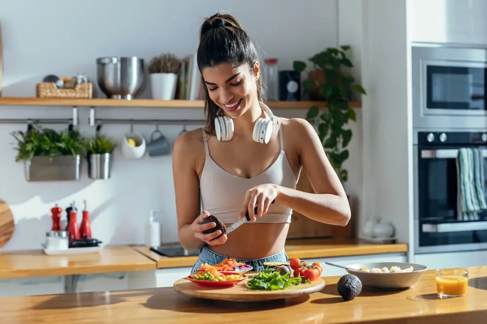 Sportlich gekleidete Frau in moderner Küche bereitet ein gesundes Gericht mit Avocado, Paprika und Blattgemüse nach dem Training zu.