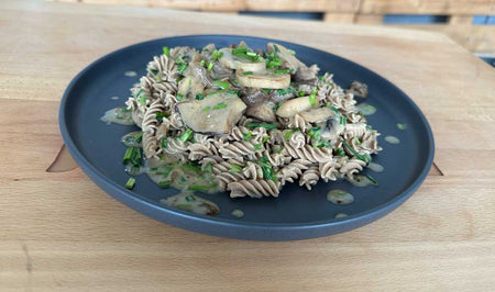 Ein Teller mit Low Carb High Protein Fusilli in Pilzrahmsoße, garniert mit frischen Kräutern und geschnittenen Frühlingszwiebeln, serviert auf einem dunkelblauen Teller auf einem Holztisch.