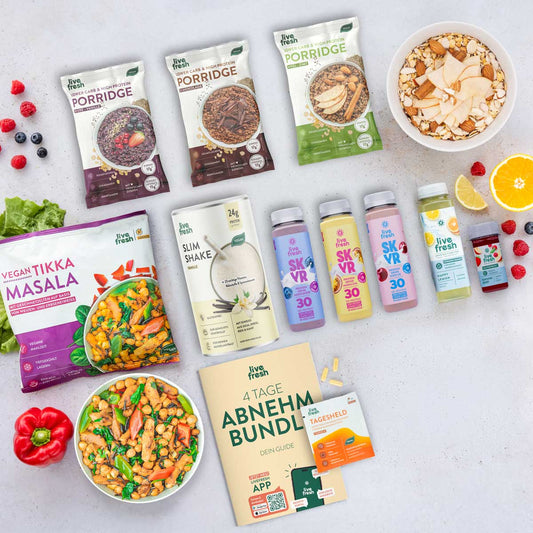 Flatlay mit dem „4 Tage Abnehm Bundle“ von LiveFresh, bestehend aus veganem Tikka Masala, Vanille Slim Shake, drei Skyr Protein Drinks (Heidelbeere, Maracuja, Kirsche), zwei Porridge-Sorten (Apfel-Zimt & Schoko), einem grünen Saft „Happy Lemon“, einem Vitamin D Shot sowie dem „Tagesheld“-Supplement. Daneben ein gedruckter Guide, frisches Obst und eine Bowl mit Nüssen und Apfelscheiben.