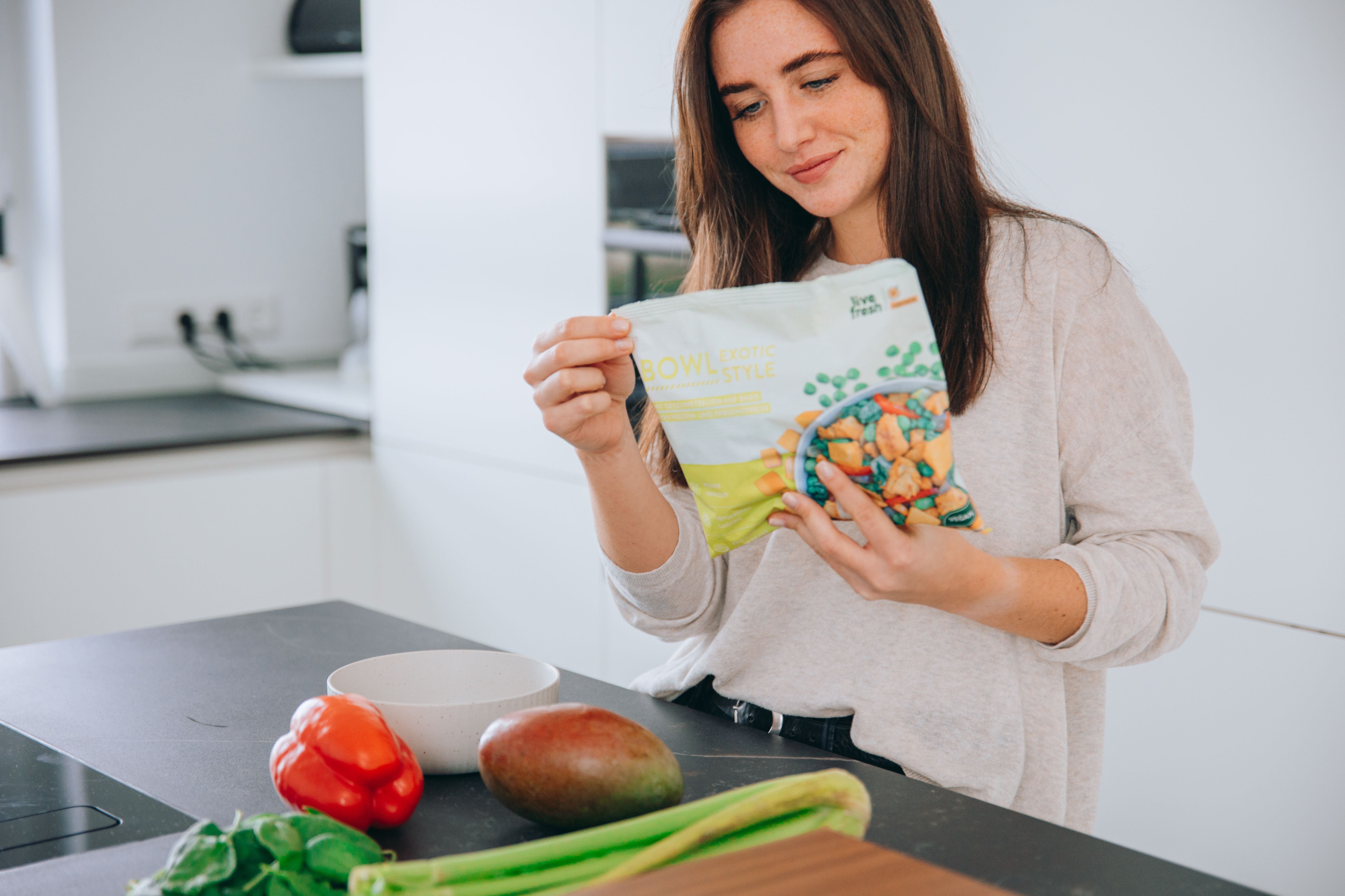 eine Frau in einer modernen Küche, die eine Packung „Live Fresh Bowl Exotic Style“ in den Händen hält und diese betrachtet. Auf der Küchenarbeitsfläche vor ihr liegen frische Zutaten wie eine rote Paprika, eine Mango, Stangensellerie und Blattgemüse. Sie bereitet sich offensichtlich auf die Zubereitung einer gesunden Mahlzeit vor, indem sie sowohl frische als auch vorbereitete Zutaten verwendet.