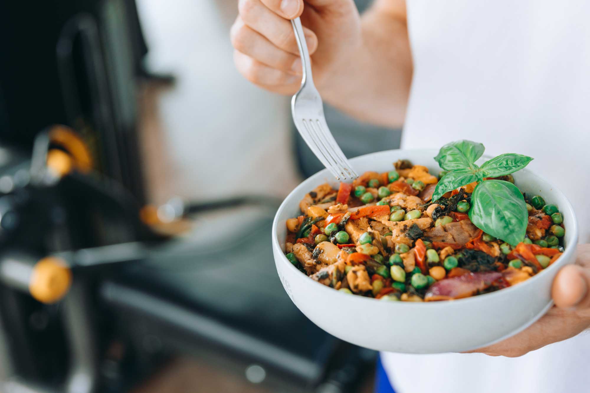 Nahaufnahme einer bunten Gemüse-Bowl mit Erbsen, Paprika, Hülsenfrüchten und frischem Basilikum, die vor einem Fitnessgerät in der Hand gehalten wird.