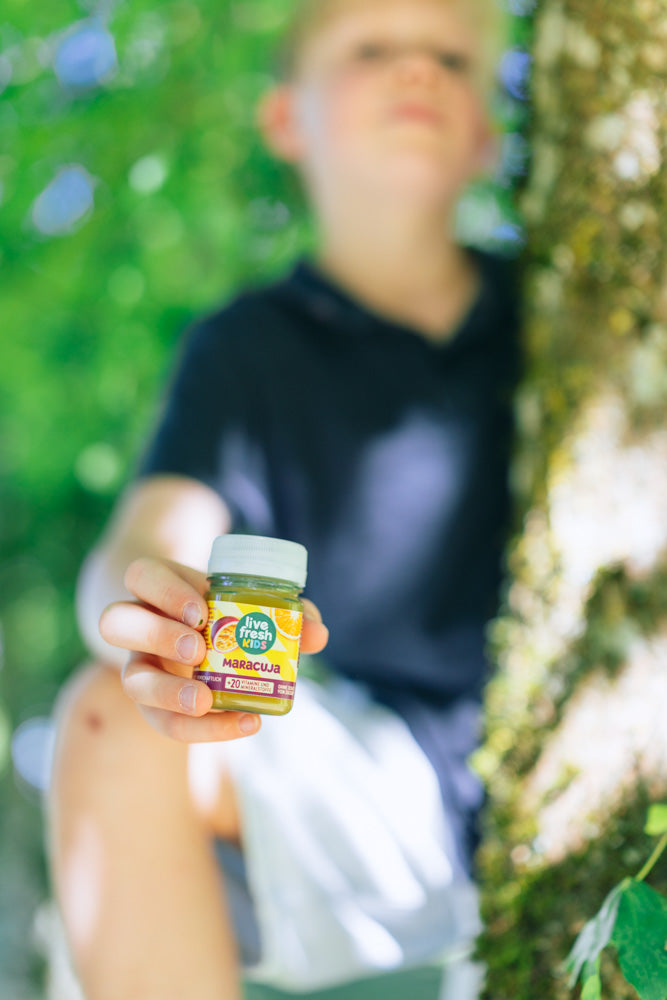 A child is sitting by a tree, holding up a LiveFresh Kids Shot Maracuja.