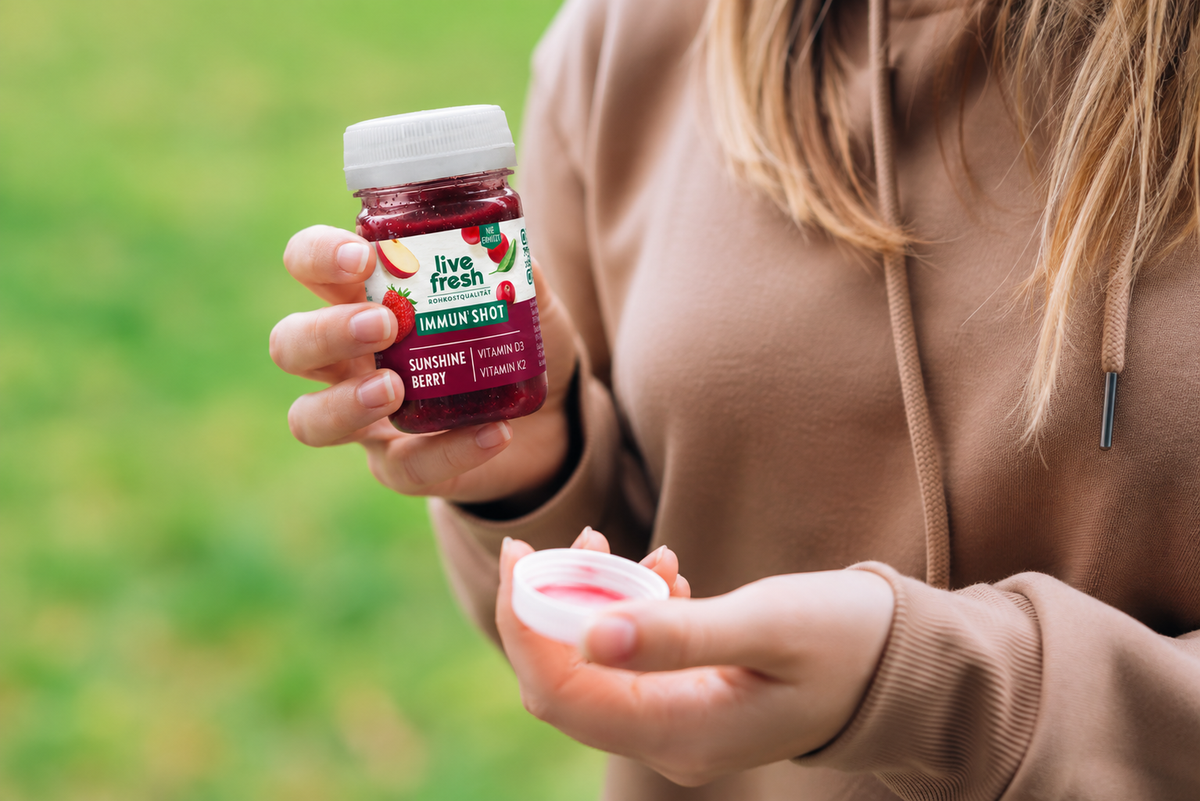 A woman is standing outside, holding the red Live Fresh Immun Shot Sunshine Berry with vitamin D3 and vitamin K2. The new compact bottle shape, with its white screw-top cap and dark red shot, is in focus against a blurred green background.