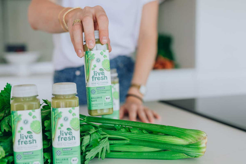 Frau greift nach einer Flasche LiveFresh Selleriesaft, die zwischen frischen Selleriestangen und weiteren Flaschen auf einer Küchenarbeitsfläche steht.