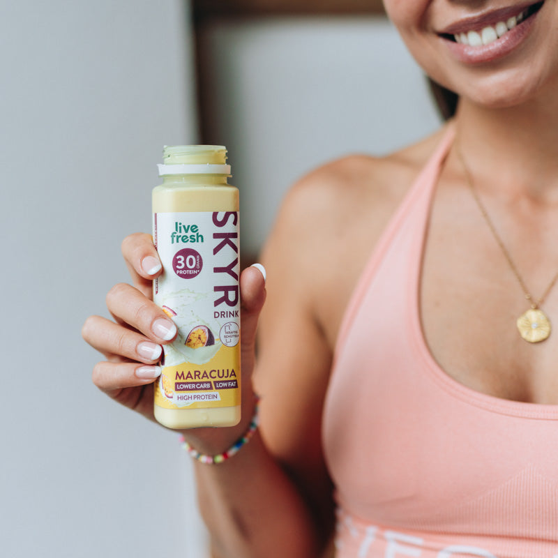 Eine Frau in einem sportlichen Top hält eine geöffnete Flasche Skyr-Drink in der Geschmacksrichtung Maracuja von 'live fresh' in der Hand und lächelt. Auf der Flasche steht '30g Protein', 'lower carb, low fat, high protein' und 'kräftig schütteln'.