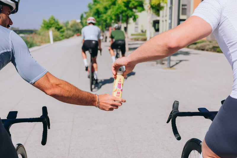 Zwei Radfahrer im Trikot übergeben sich während der Fahrt eine Flasche Skyr-Drink von LiveFresh auf einem sonnigen Stadtweg mit Bäumen im Hintergrund.