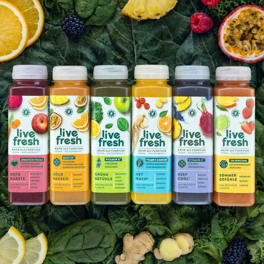Sechs verschiedene „LiveFresh“ Säfte, die nebeneinander auf grünem Blattgemüse angeordnet sind. Die Sorten umfassen „Rote Rakete“, „Goldrausch“, „Grüne Gefühle“, „Hey Wach!“, „Keep Cool!“ und „Sommergefühle“. Jede Flasche hebt spezifische Gesundheitsvorteile hervor, wie Vitamin C, Biotin oder Koffein. Um die Flaschen herum liegen frische Zutaten wie Orangen, Ingwer, Ananas und Beeren, die die Frische und die natürlichen Inhaltsstoffe der Säfte betonen.