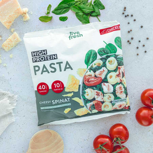 Das Bild zeigt eine Packung „Live Fresh High Protein Pasta“ in der Variante Cheesy Spinat mit Hähnchenbrustfilet. Die Verpackung hebt den hohen Proteingehalt von 40 g und eine Zubereitungszeit von 10 Minuten hervor. Frische Zutaten wie Spinatblätter, Kirschtomaten und Käse sind um die Packung verteilt, was die cremige und proteinreiche Pasta unterstreicht.