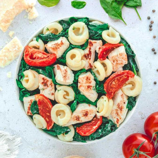 Ein weiteres Bild zeigt die angerichtete Pasta-Schüssel mit gedrehten Nudeln, Hähnchenstücken, Spinatblättern und halbierten Tomaten, die auf frischem Blattspinat liegen. Die um den Teller platzierten Zutaten wie Käsewürfel, Spinat und Tomaten runden das farbenfrohe Gericht ab und betonen die hochwertige Zubereitung.