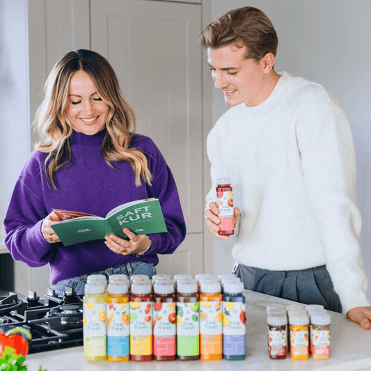 Eine Frau im lila Pullover und ein Mann im weißen Pullover stehen in einer modernen Küche und schauen gemeinsam in ein grünes „SAFT KUR“-Heft. Der Mann hält eine Flasche Live Fresh Saft in der Hand. Vor ihnen auf der Kücheninsel stehen mehrere farbenfrohe Säfte und Shots in einer Reihe – das sogenannte „Couple Friends Bundle“.