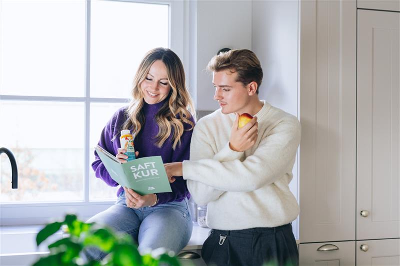Zwei Personen in einer hellen Küche, die gemeinsam in einen "Saftkur"-Guide von LiveFresh schauen, während die Frau einen Saft hält und der Mann einen Apfel isst.