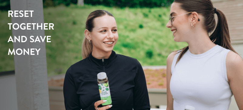 Two women in fitness clothing smile at each other while the one on the left with a black vest, holds a bottle of juice called Grüne Gefühle by LiveFresh. On the left upper side of the picture it says 