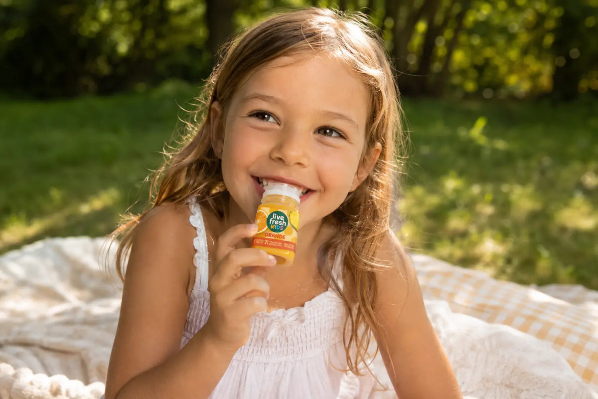 Ein Mädchen auf einer Picknickdecke im Grünen trinkt lächelnd einen LiveFresh Kids Orangensaft. 