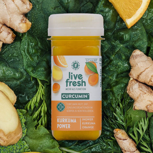 LiveFresh Shot „Kurkuma Power“ mit Curcumin für entzündungshemmende Wirkung, leuchtend gelb-orange. Umgeben von Kurkuma, Ingwer und Orangenscheiben auf grünem Blattgemüse.