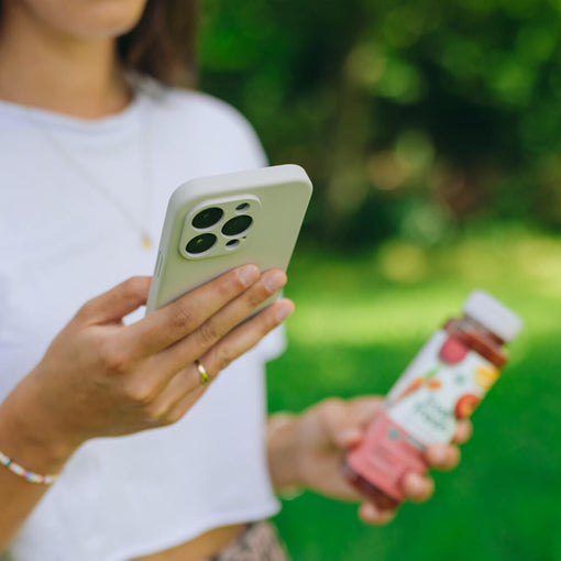Eine Frau im weißen T-Shirt hält in der linken Hand ein Smartphone mit heller Hülle und in der rechten eine Flasche LiveFresh Saft. Der Fokus liegt auf dem Handy, während der Hintergrund mit grünem Rasen und Bäumen verschwommen ist – die Szene vermittelt einen modernen, gesunden Lifestyle unterwegs, wobei die LiveFresh App unterstützen soll.