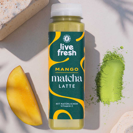 Flasche eines Mango Matcha Latte steht auf hellem Untergrund neben einer frischen Mangospalte und einem Haufen grünem Matchapulver. Auf dem Etikett steht "live fresh mango matcha latte mit natürlichem vitamin c" in modernem Design mit grünen und gelben Elementen. Die Kombination aus Frucht und Pulver verdeutlicht die Zutaten und den frischen Charakter des Getränks.