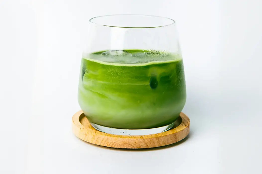 Ein bauchiges Glas mit grünem Matcha Latte auf einem Holz Untersetzer vor hellgrauem Hintergrund