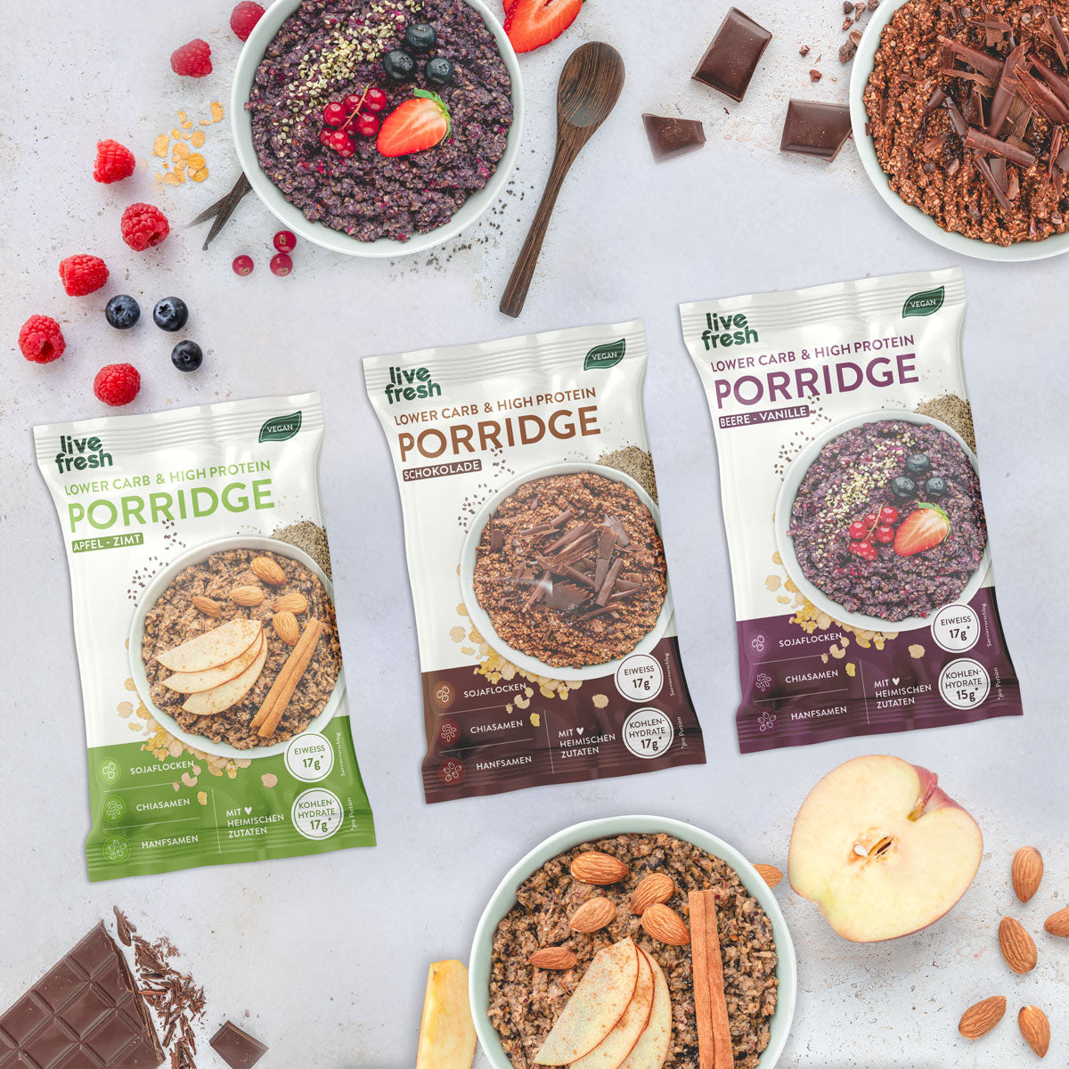 Zeigt drei Packungen „Live Fresh“ Porridge in den Sorten Apfel-Zimt, Schokolade und Beere-Vanille. Jede Packung ist als Lower Carb & High Protein gekennzeichnet und enthält Sojaflocken, Chiasamen und Hanfsamen. Um die Packungen herum sind Schalen mit den zubereiteten Porridges, dekoriert mit frischen Früchten, Schokolade und Zimtstangen, sowie einige Zutaten wie Beeren, Apfelstücke und Mandeln verteilt.