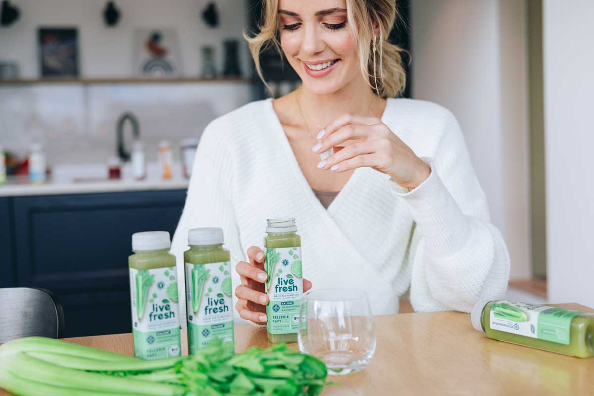 Eine Frau sitzt an einem Tisch und hält eine Flasche Live Fresh Selleriesaft in der Hand, während mehrere weitere Flaschen des Safts auf dem Tisch stehen. Vor ihr liegen frische Selleriestangen und ein leeres Glas.