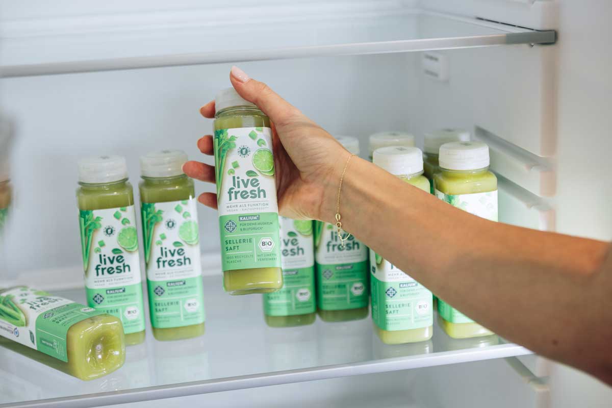 Eine Hand greift nach einer Flasche Live Fresh Selleriesaft aus einem Kühlschrank, in dem weitere Flaschen desselben Safts stehen. Die Szene wirkt ordentlich und kühl, was den frischen und gesunden Charakter des Produkts unterstreicht. 