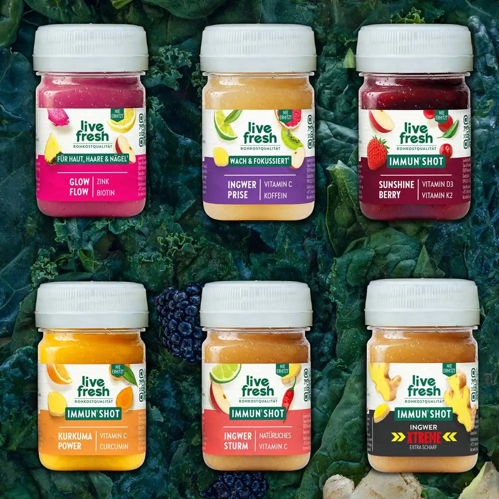 Sechs Live Fresh Shots sind in zwei Reihen auf dunkelgrünem Blattgemüse angeordnet. Zu sehen sind die Sorten Glow Flow, Ingwer Prise, Sunshine Berry, Kurkuma Power, Ingwer Sturm und Ingwer Xtreme mit unterschiedlichen Vitaminen und Inhaltsstoffen.