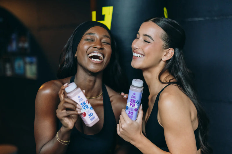 Zwei lachende Frauen in schwarzer Sportkleidung halten LiveFresh SK VR Protein Drinks mit 30 Gramm Protein in der Hand vor dunklem Hintergrund.