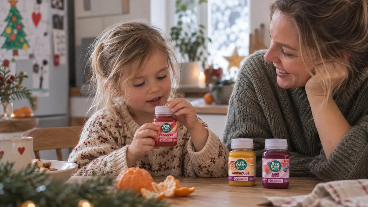 Ein Mädchen hält am weihnachtlich dekorierten Küchentisch eine Flasche LiveFresh Kids Erdbeere in den Händen während eine Frau lächelnd zusieht. Daneben stehen die Sorten Maracuja und Waldbeere sowie eine geschälte Mandarine.