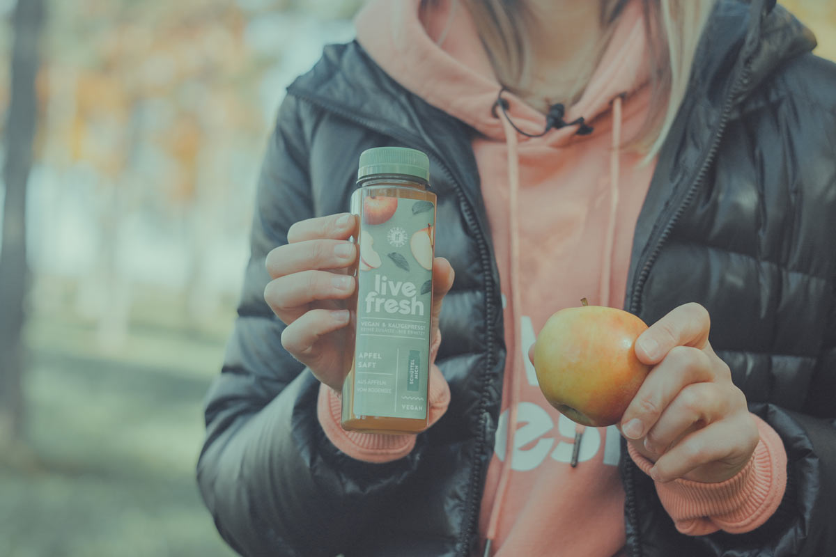 Eine Person in einer schwarzen Jacke und einem rosafarbenen Hoodie, die eine Flasche „Live Fresh“ Apfelsaft in der einen Hand und einen frischen Apfel in der anderen hält. Auf der Saftflasche steht „Vegan & Kaltgepresst“, sowie „Apfelsaft mit Äpfeln vom Bodensee“. Der Hintergrund zeigt eine unscharfe, herbstliche Umgebung im Freien.